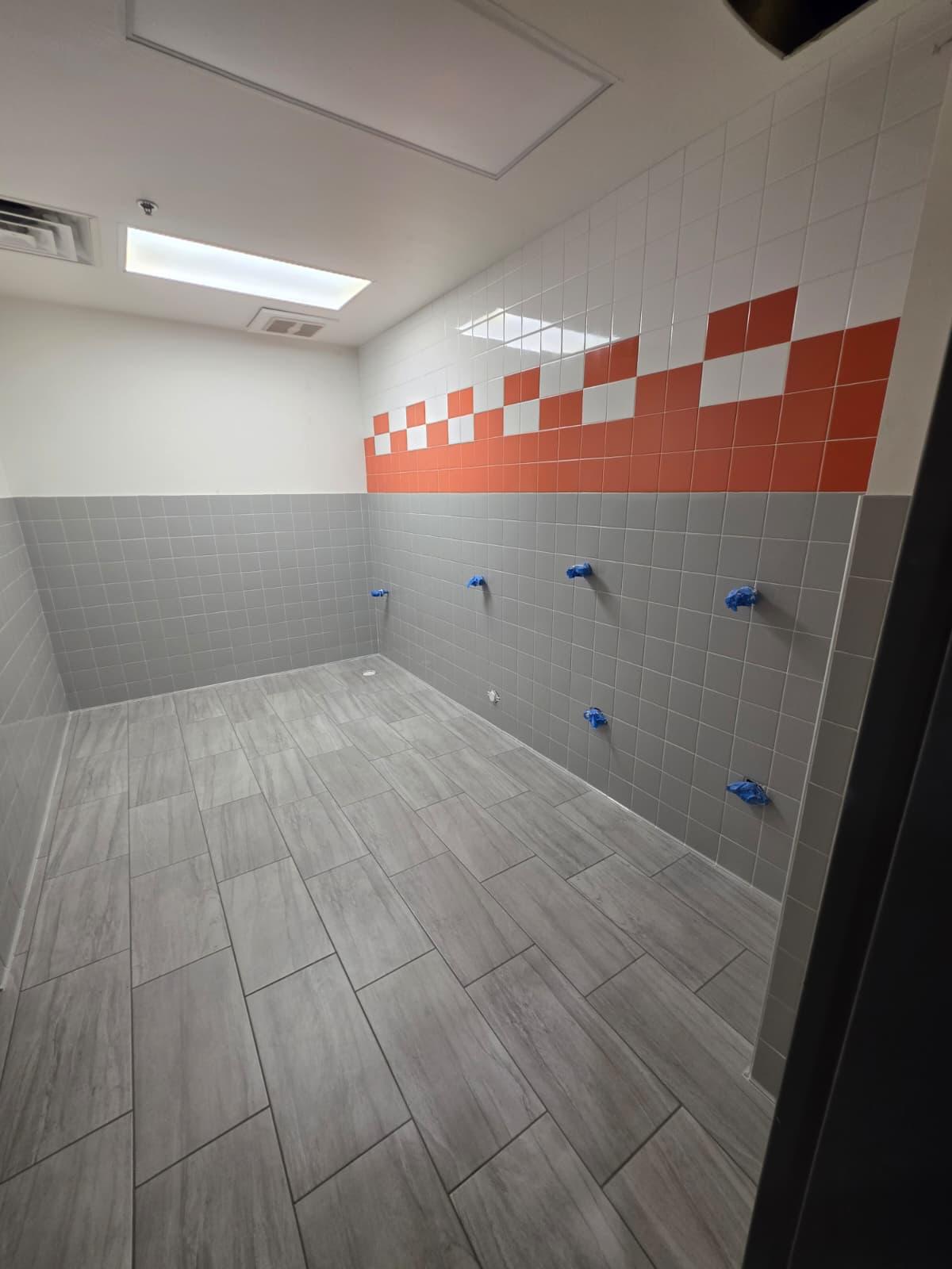 Wall and floor tile in a commercial wash room — grey field tile with an orange accent band and wood-look plank floor tile.